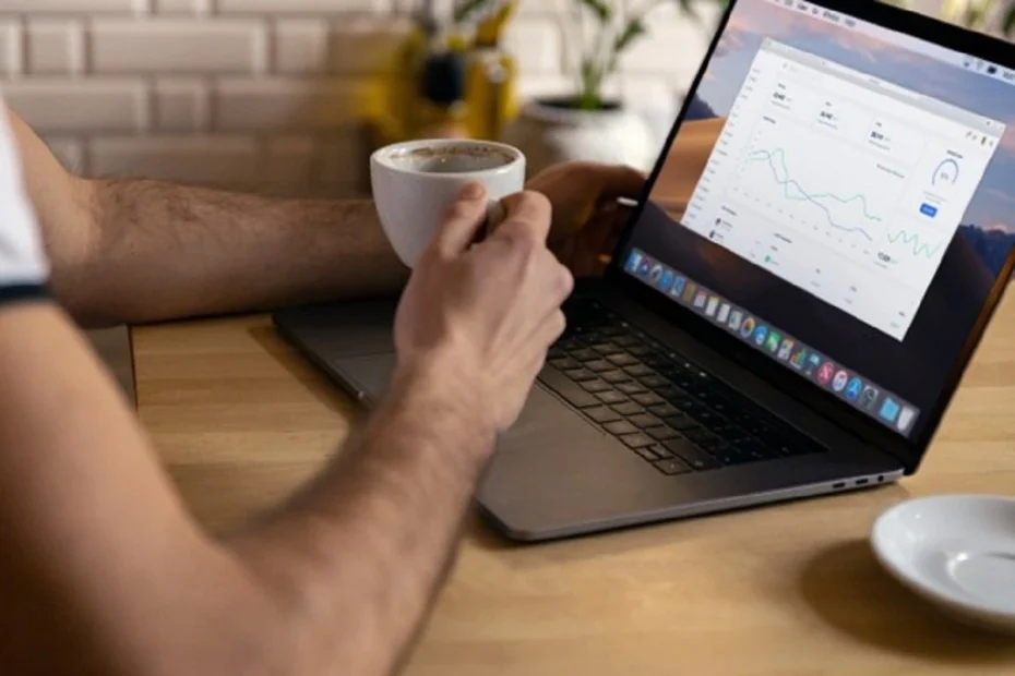 Person holding a coffee cup while working on a laptop with charts, representing making money from home.